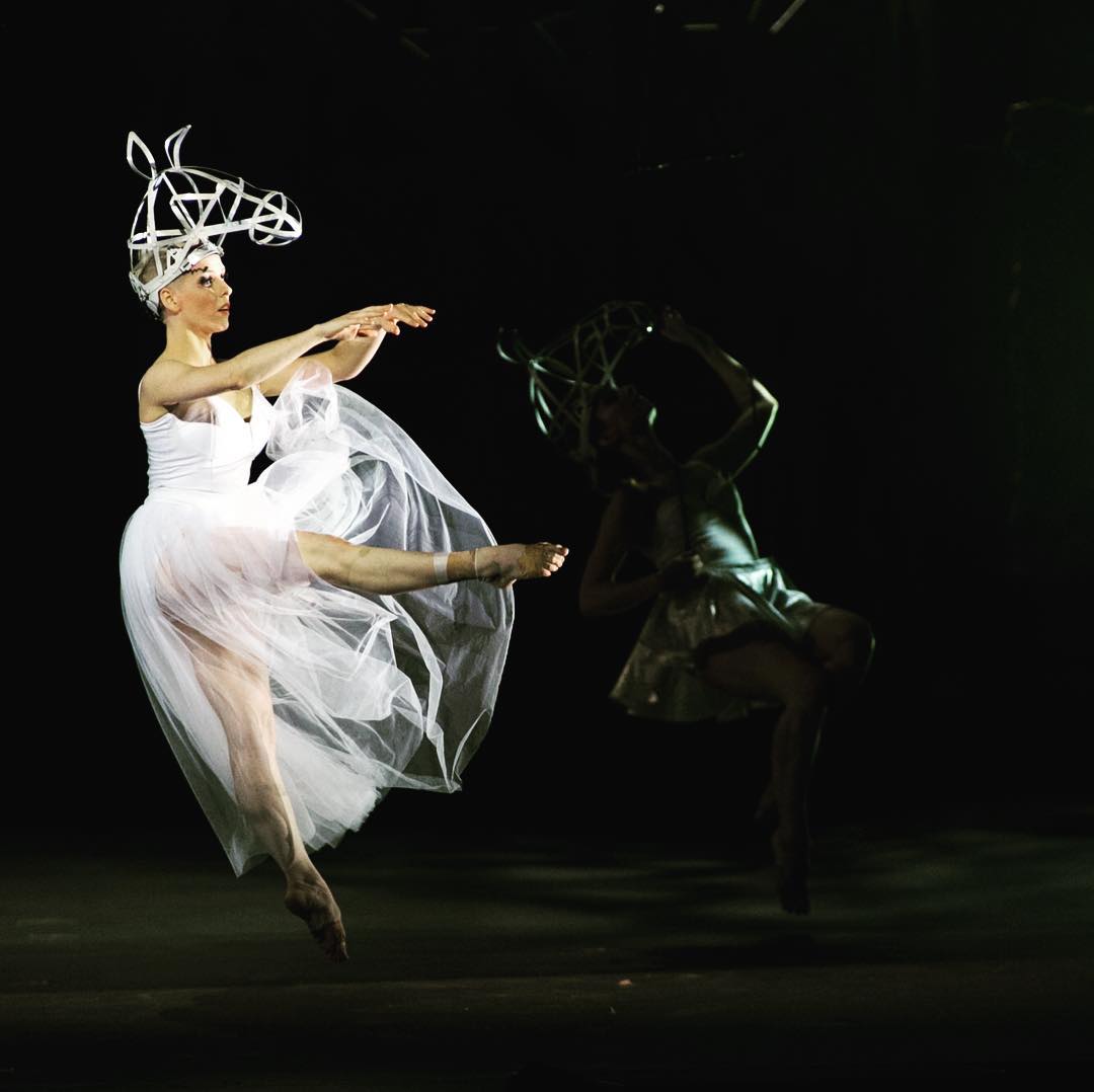 On stage in a white tulle dress, Merryn is jumping, arms and one leg extended in front of her.